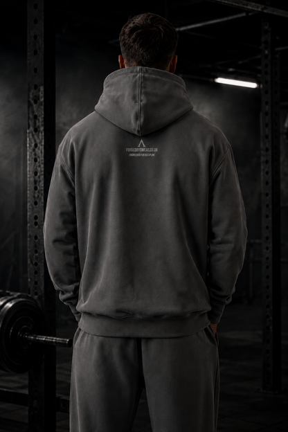 A man viewed from behind wearing a dark gray hoodie and matching sweatpants in a dimly lit gym setting. A small white logo is printed on the upper back of the hoodie.
