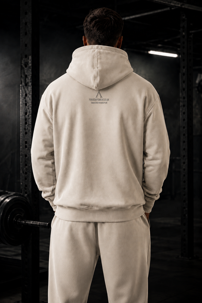 A man viewed from behind wearing a light beige hooded sweatshirt and matching sweatpants in a dark gym setting.